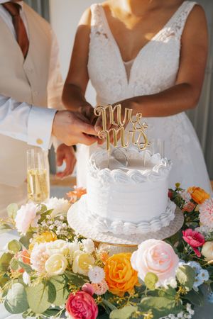 White wedding cake with gold Mr and Mrs topper surrounded by colorful roses and greenery at reception.