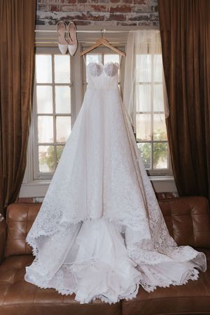 Elegant white lace wedding dress with sweetheart neckline hangs from wooden hanger against brown curtains and leather couch.
