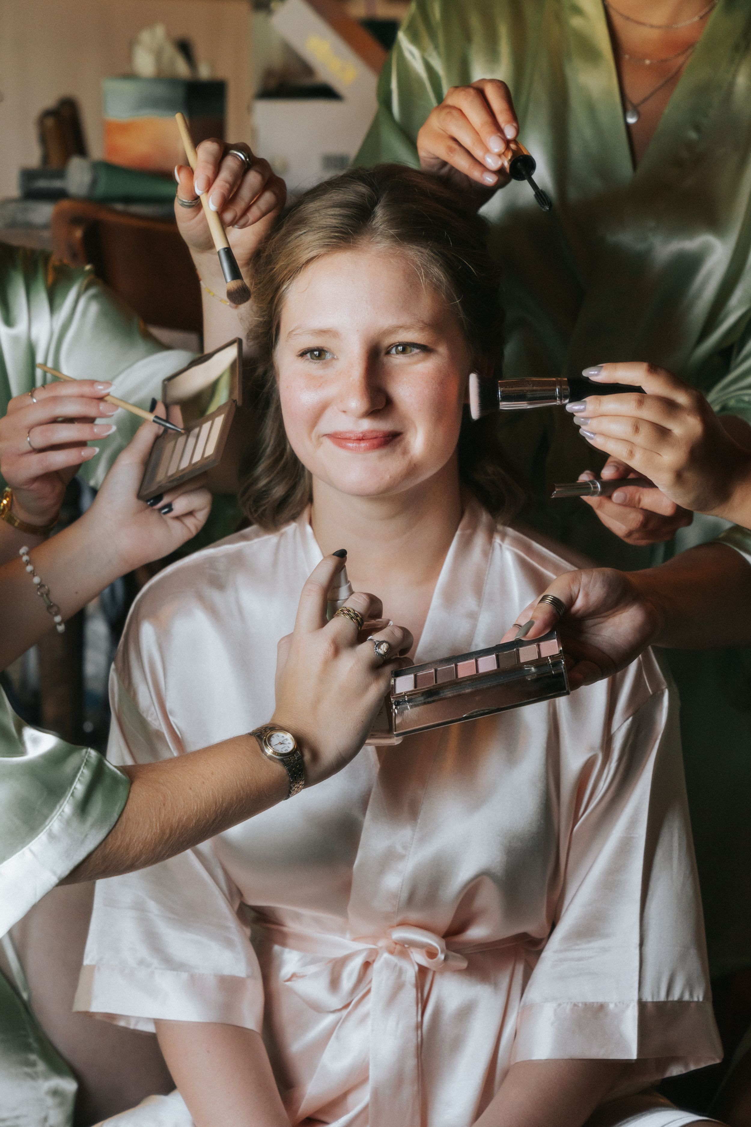 Multiple hands apply makeup with brushes and tools during a beauty preparation session.