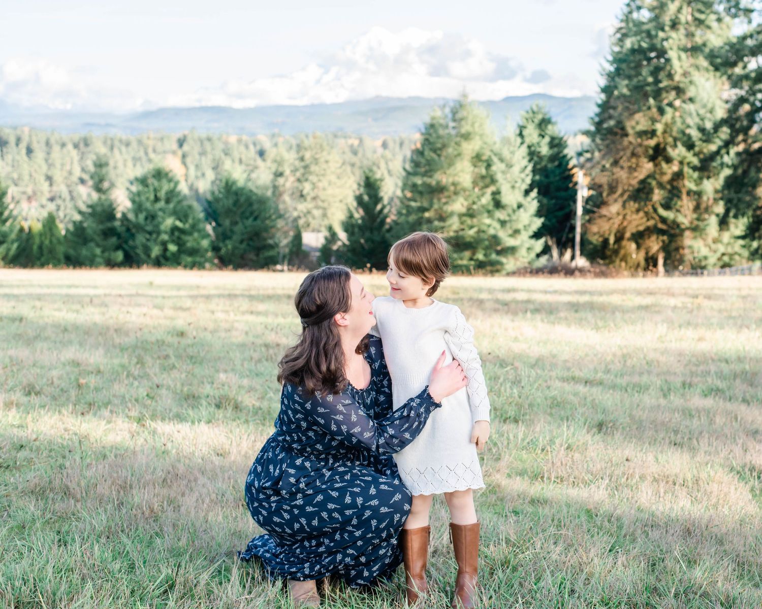 Two figures share a sweet moment in a grassy meadow with evergreen forest and mountains in the background.