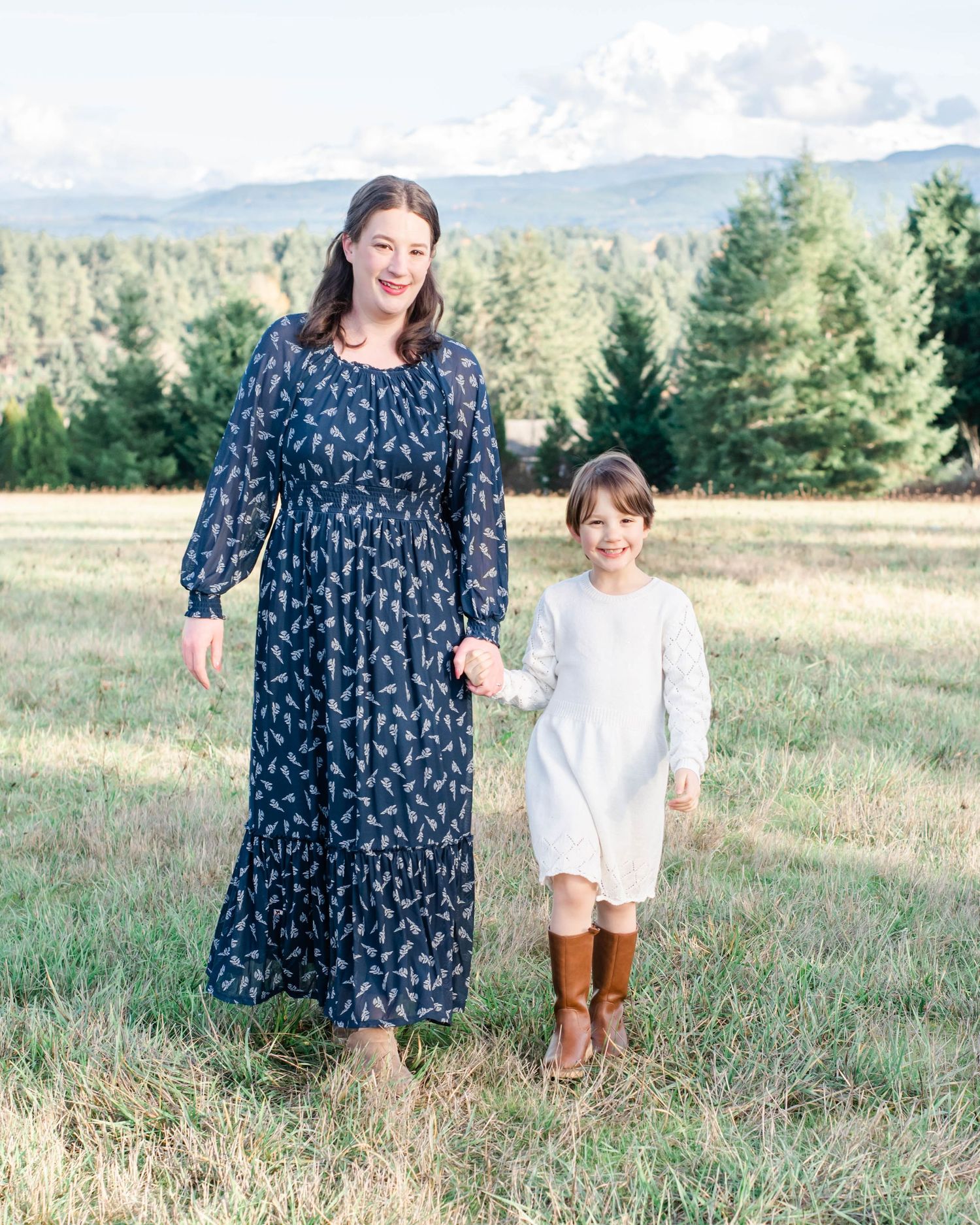 A pair stands together in a floral maxi dress and white dress against a mountain landscape.