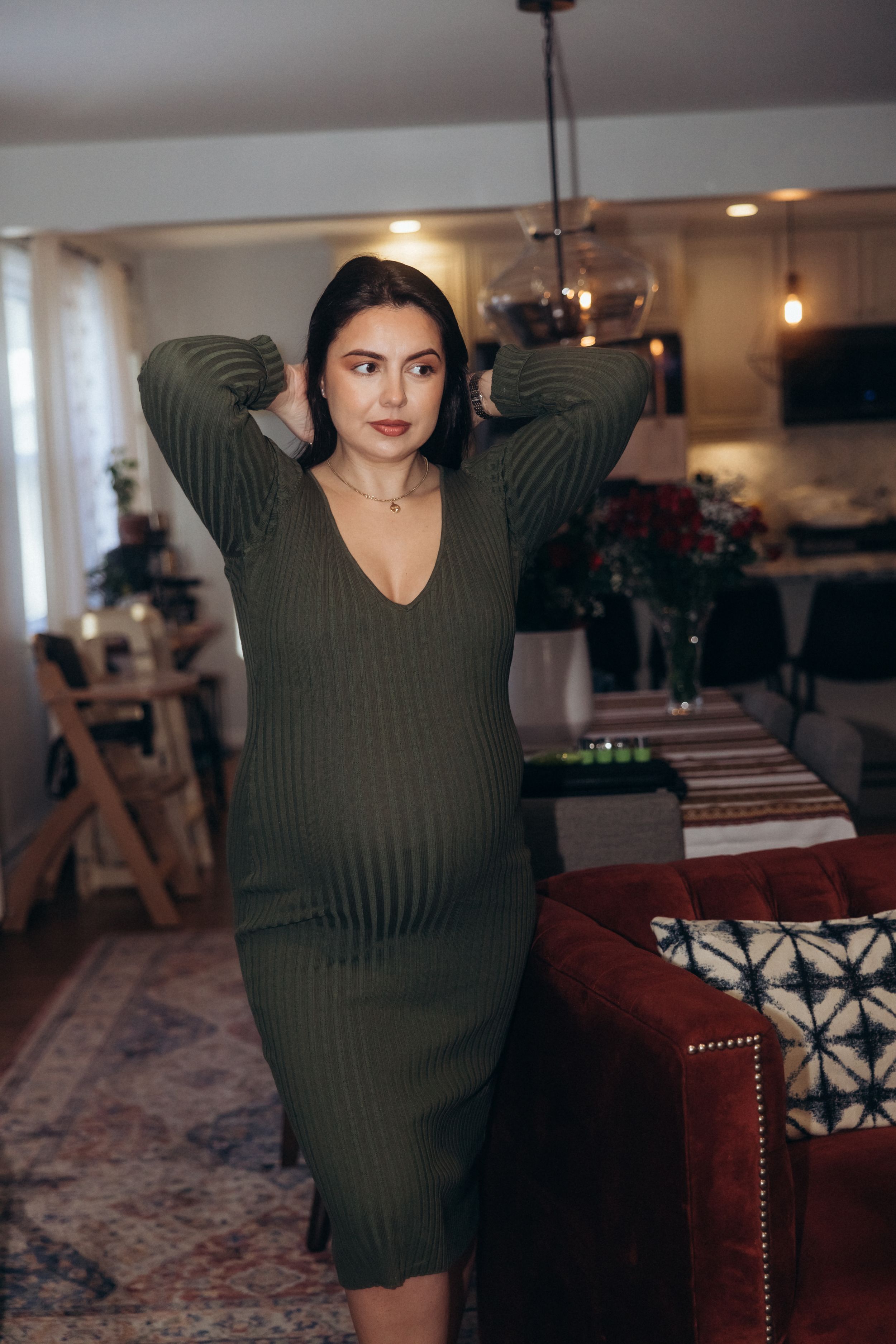 A pregnant woman in a dark green ribbed dress poses in a modern living room setting.