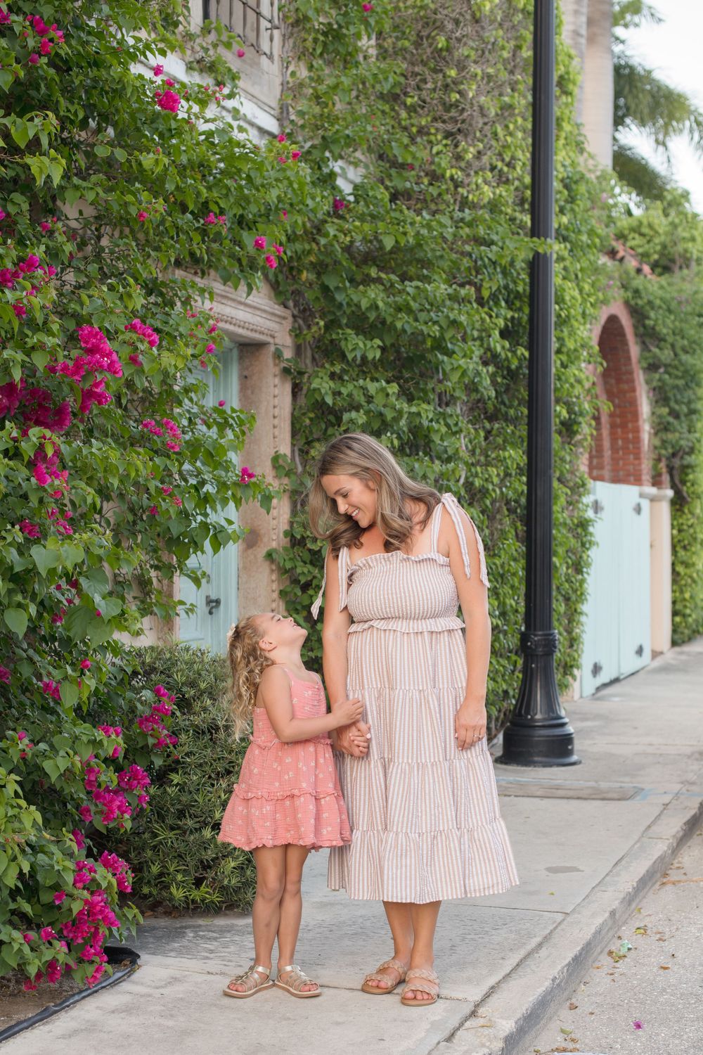 Mother and daughter in summer dresses walking along path with flowering vines.