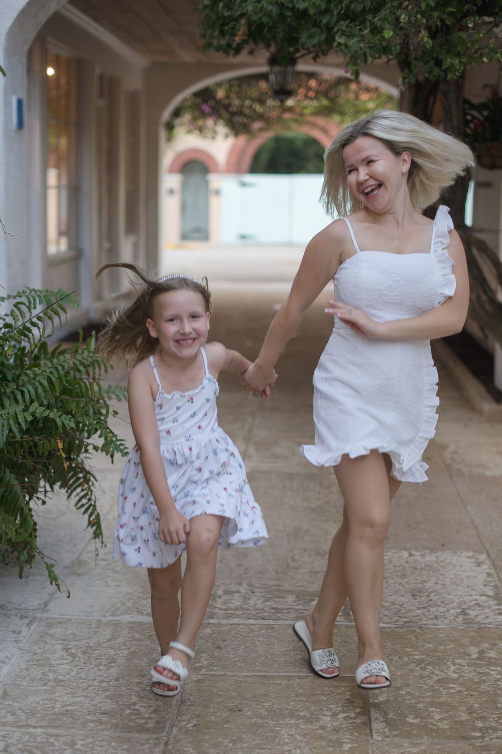 Mother and daughter in white outfits playing together under covered walkway.