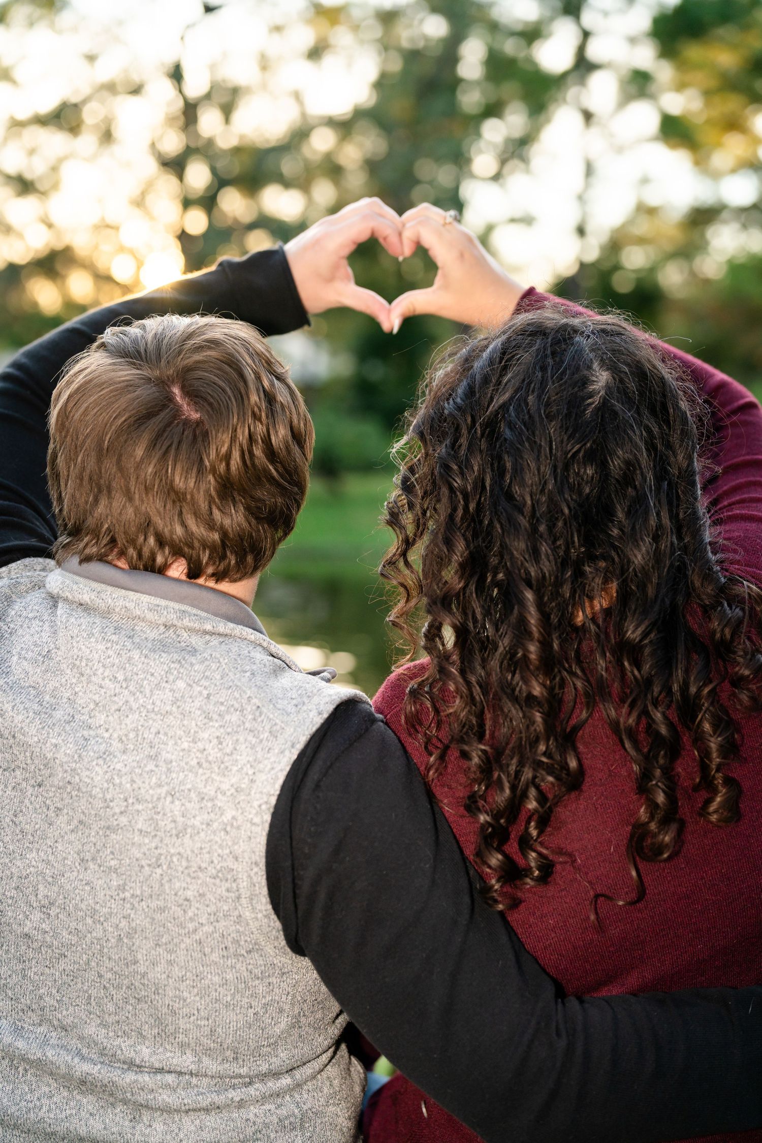 Two individuals forming a heart shape with their hands against a blurred natural background at sunset.