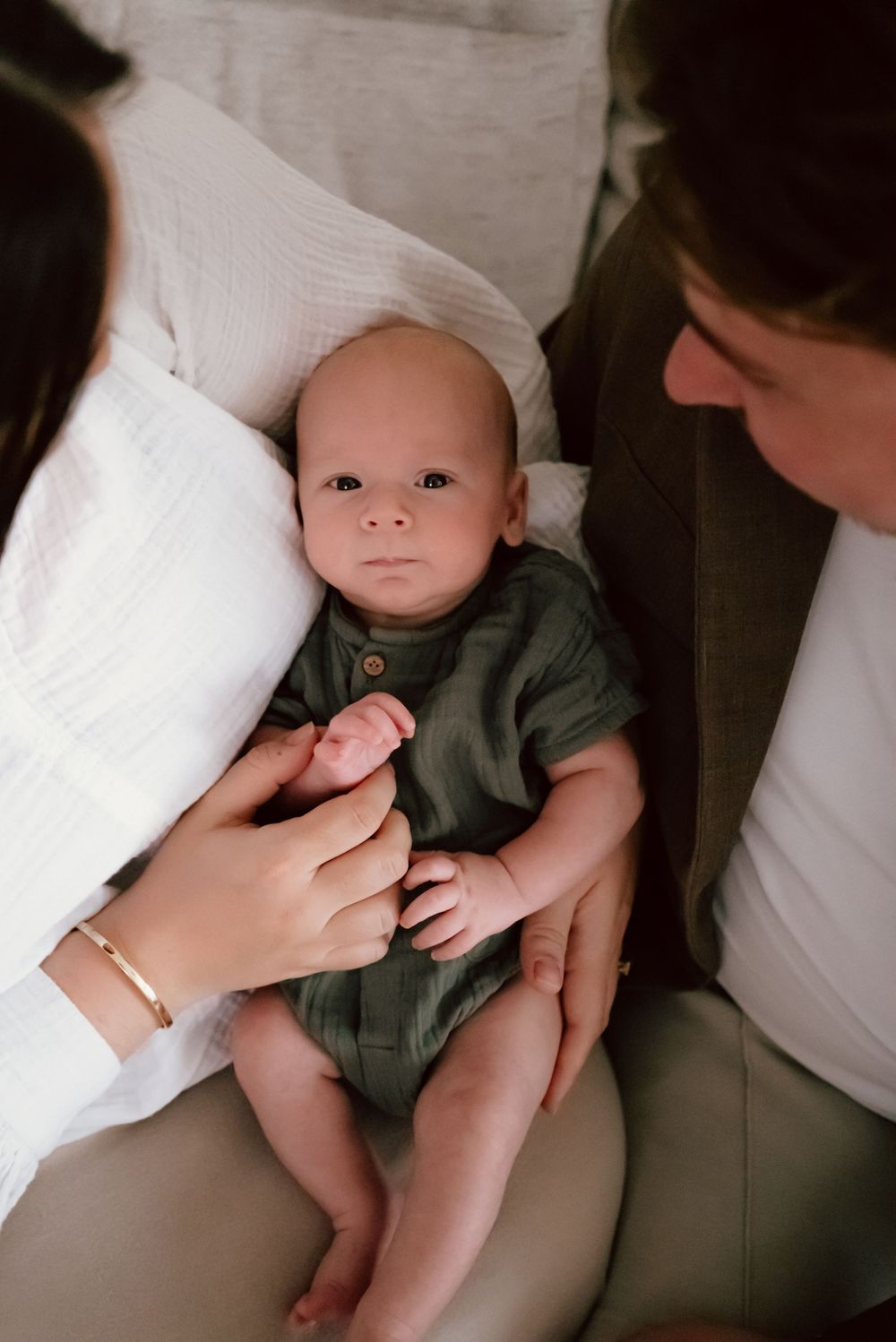 A baby wearing a green onesie lies on white bedding while being lovingly touched by caring hands.