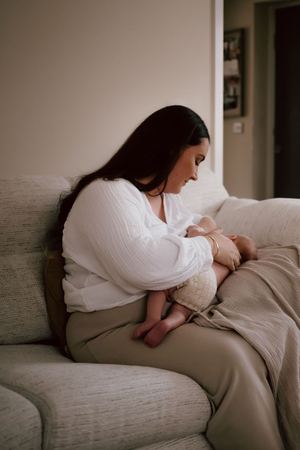 A person in white attire cradles and nurses a baby while seated on a gray couch in a softly lit room.