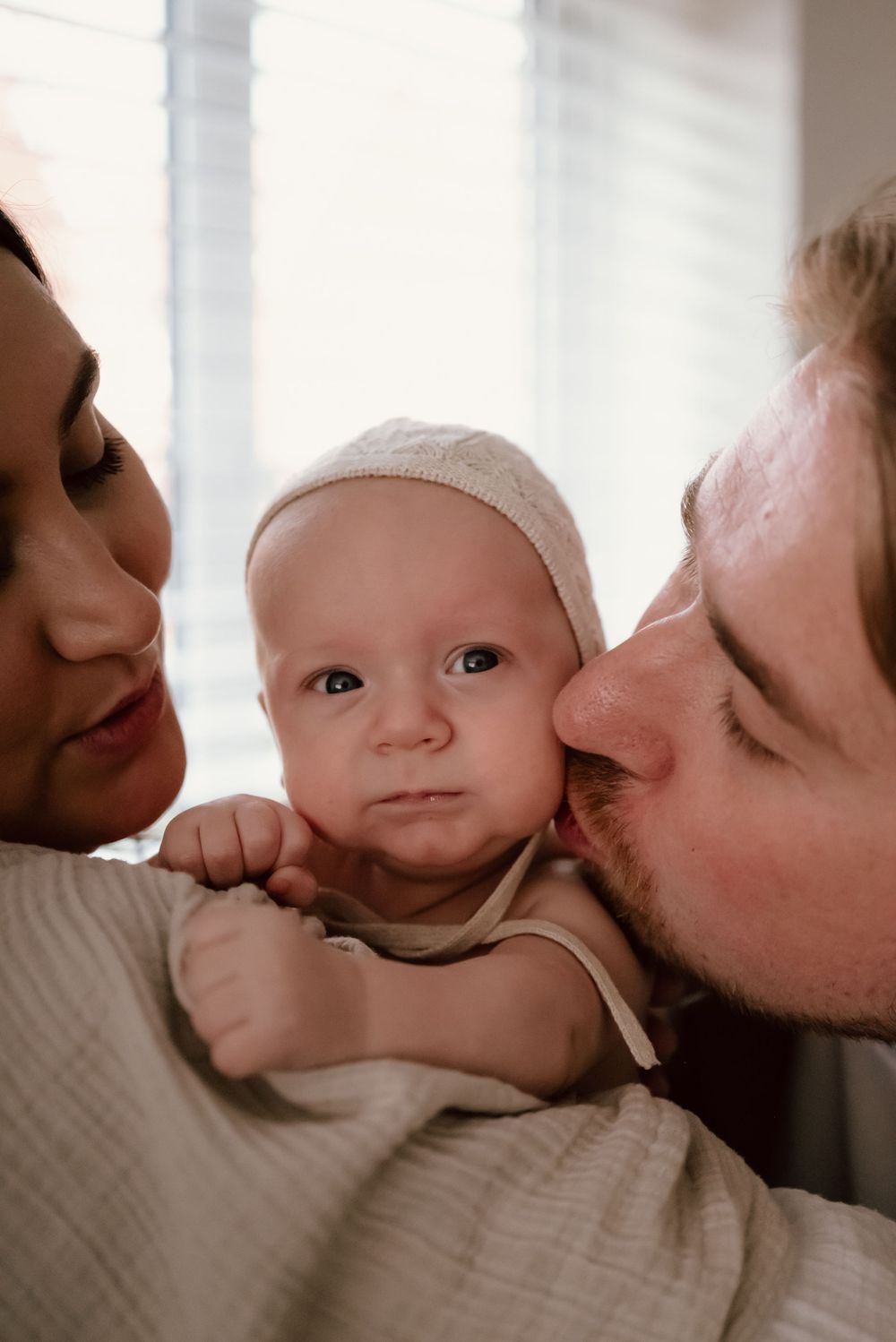 An intimate family portrait shows parents lovingly embracing their baby wearing a white cap in soft natural lighting.