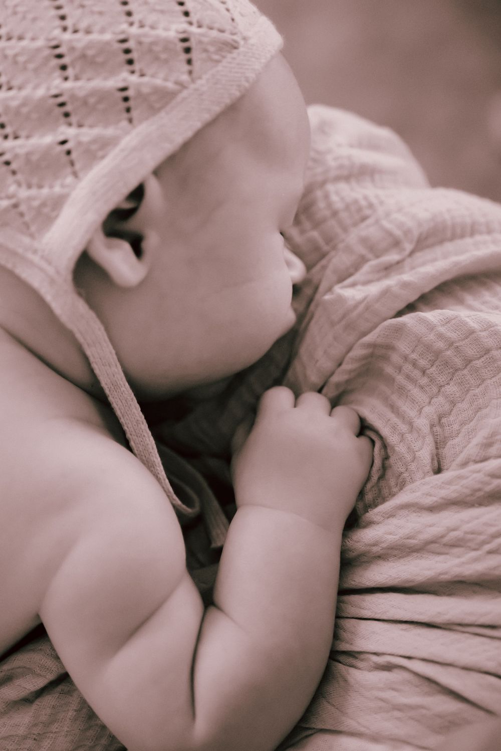 A sleeping infant in soft sepia tones wearing a knit bonnet and swaddled in textured fabric.