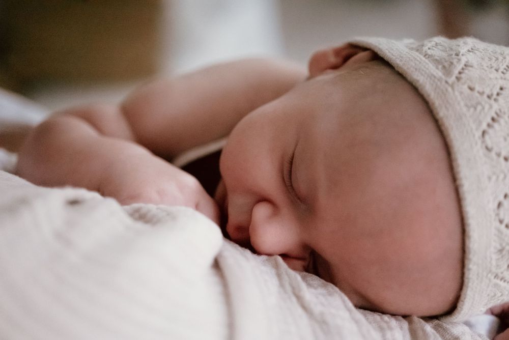 A peacefully sleeping newborn baby wearing a delicate white lace bonnet rests against soft white fabric.