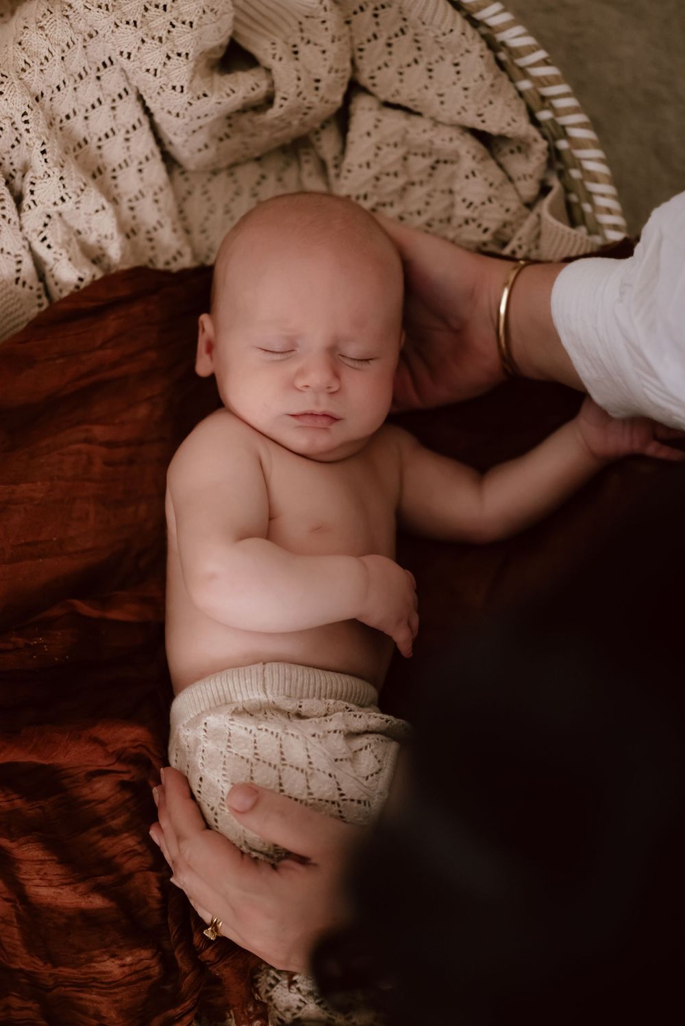Sleeping newborn lying on dark fabric wearing a knitted nappy cover while someone's hand gently rests nearby.