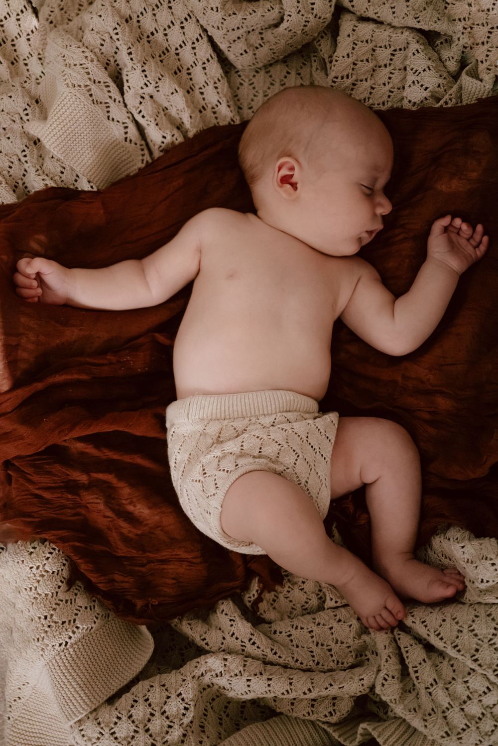 A sleeping infant wearing a knit bloomers cover lies peacefully on brown velvet fabric with cream lace blankets.