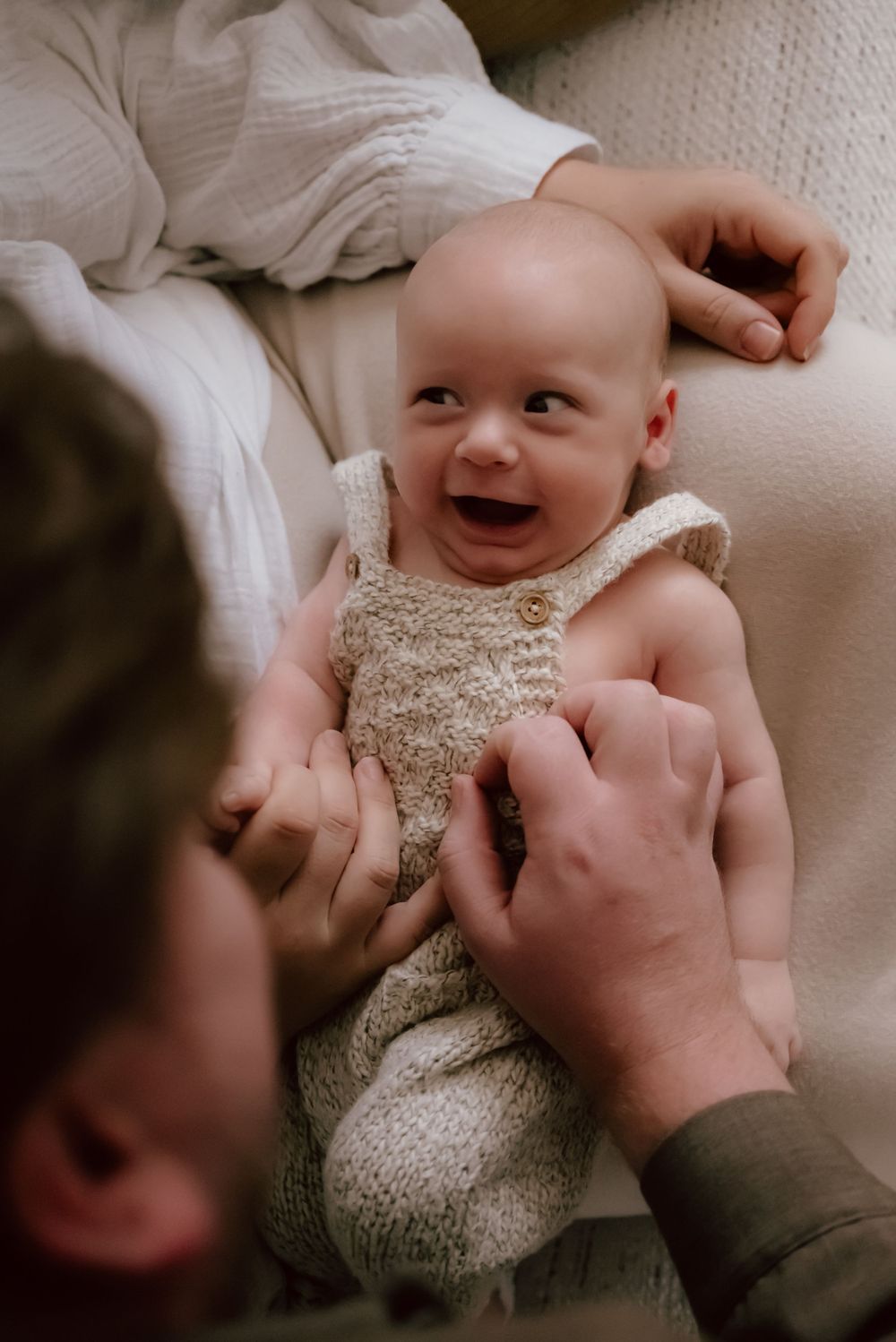 A joyful baby wearing a beige knit romper lies on a white surface while hands reach to tickle their tummy.
