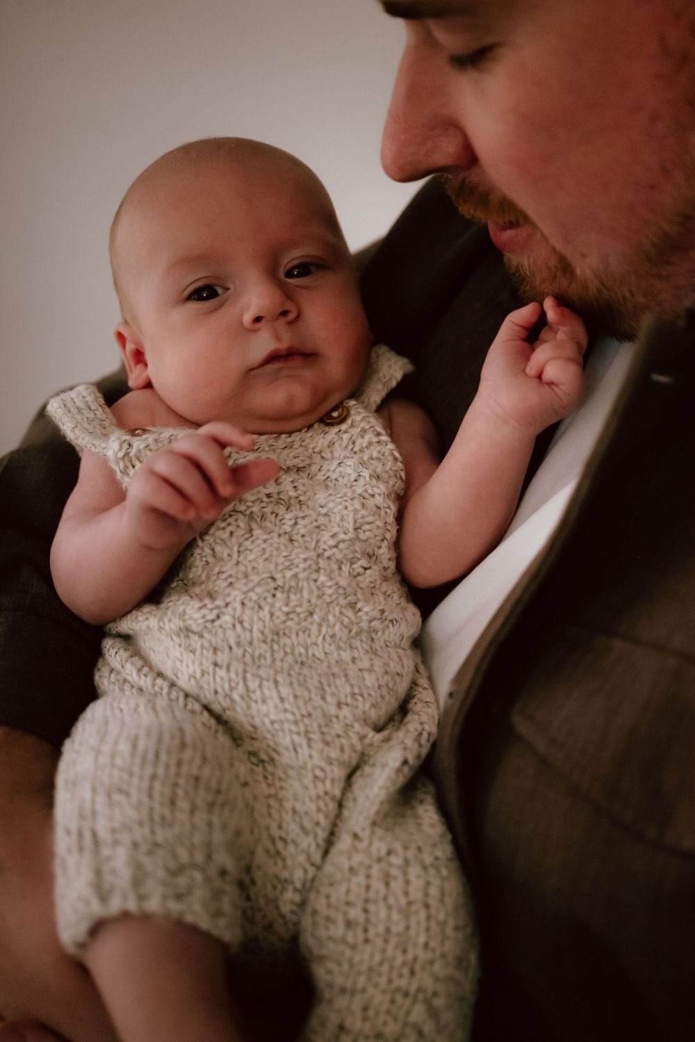 A baby wearing a knit cream-colored romper is held close while resting in loving arms.