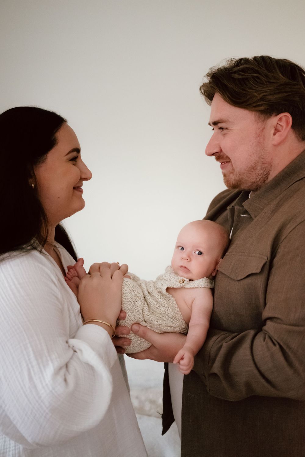 Young couple shares a tender moment while holding their small baby in a neutral-toned indoor setting.