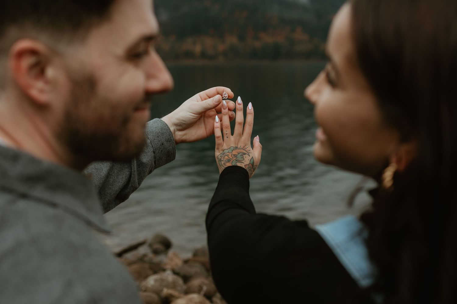 A couple holds hands by a lake showing off an engagement ring.