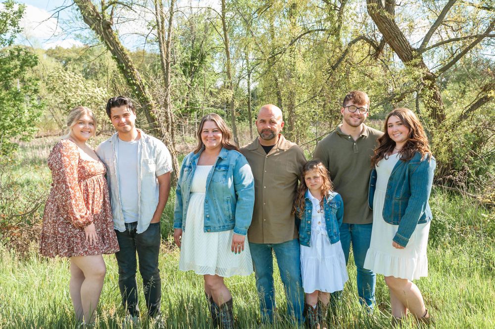 Outdoor family portrait with six people in casual attire standing together against a natural wooded backdrop.
