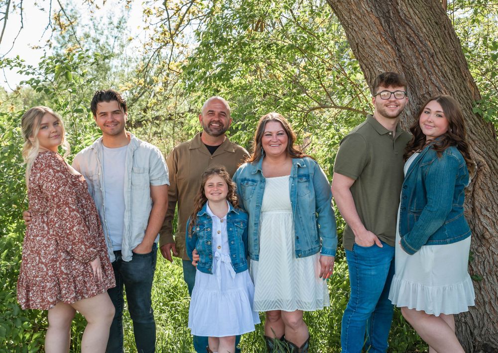 A casual family portrait outdoors with seven people dressed in coordinating blue and neutral tones.