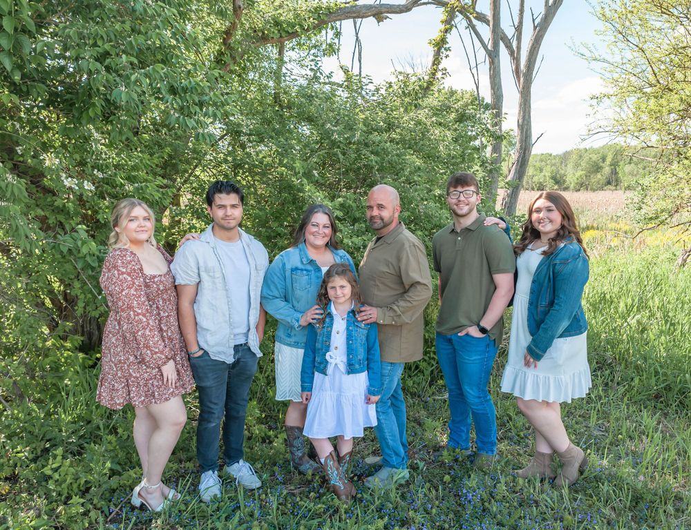 Seven people pose for a casual family photo in a natural outdoor setting with green foliage.