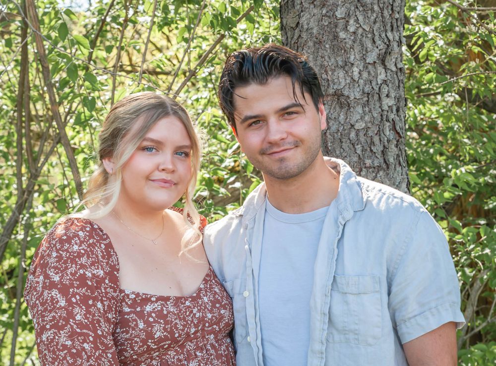 A couple poses together in front of a tree wearing casual attire.