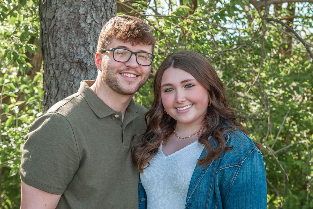 A couple smiles for a portrait against a leafy green background.