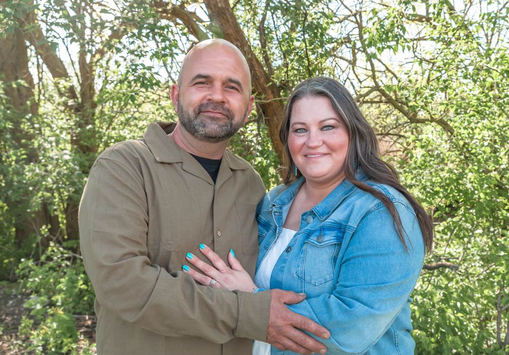 Couple in casual blue and brown clothing embracing outdoors with green foliage in background.