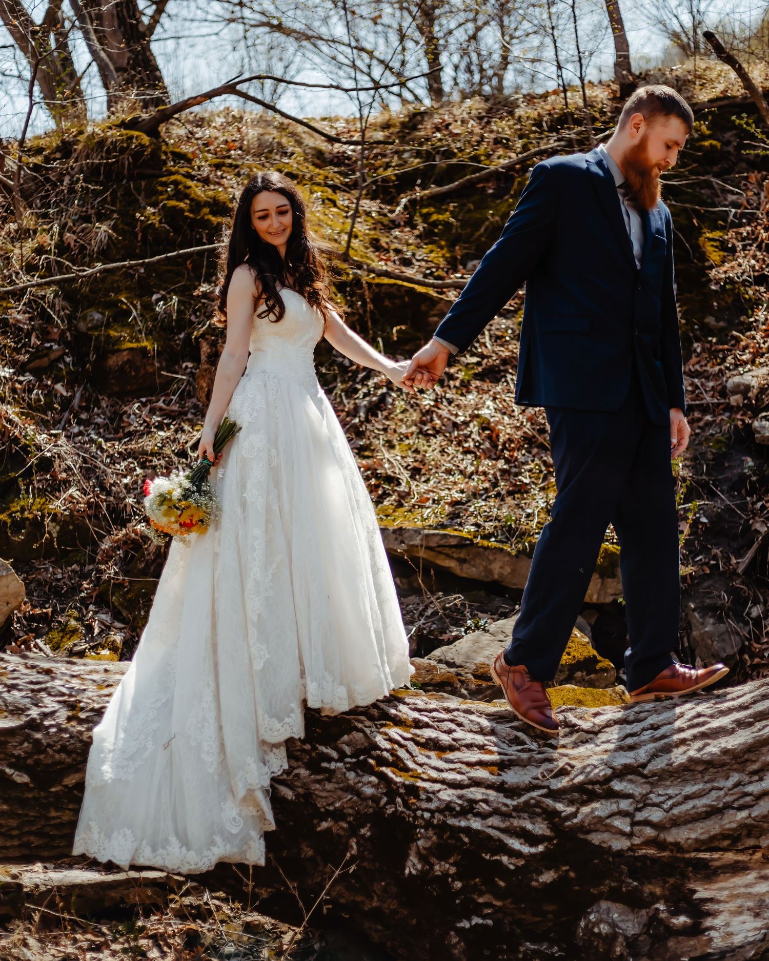 Couple holding hands while walking across rocky stream in wedding attire.