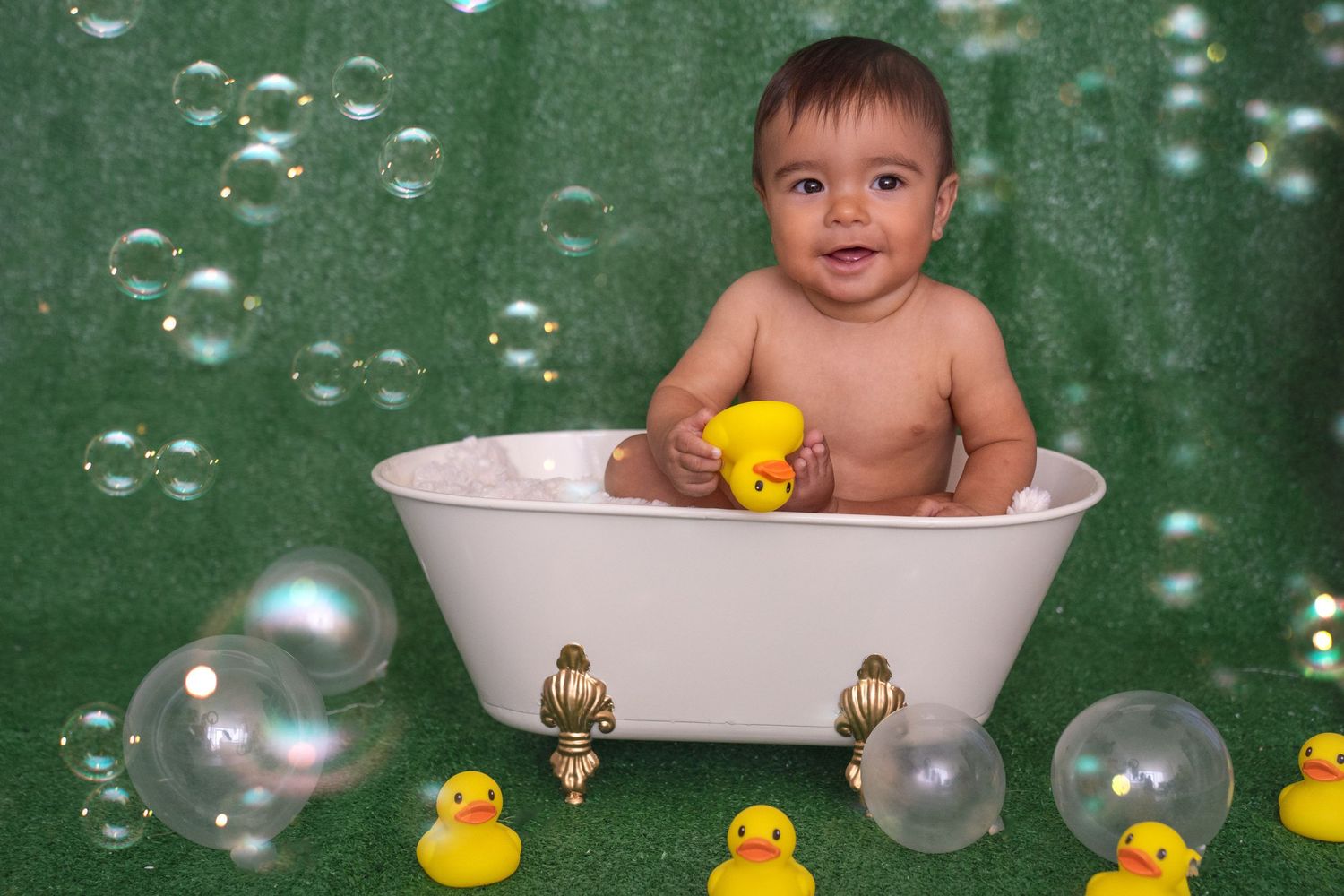 Baby in bathtub with rubber ducks and soap bubbles against green backdrop.
