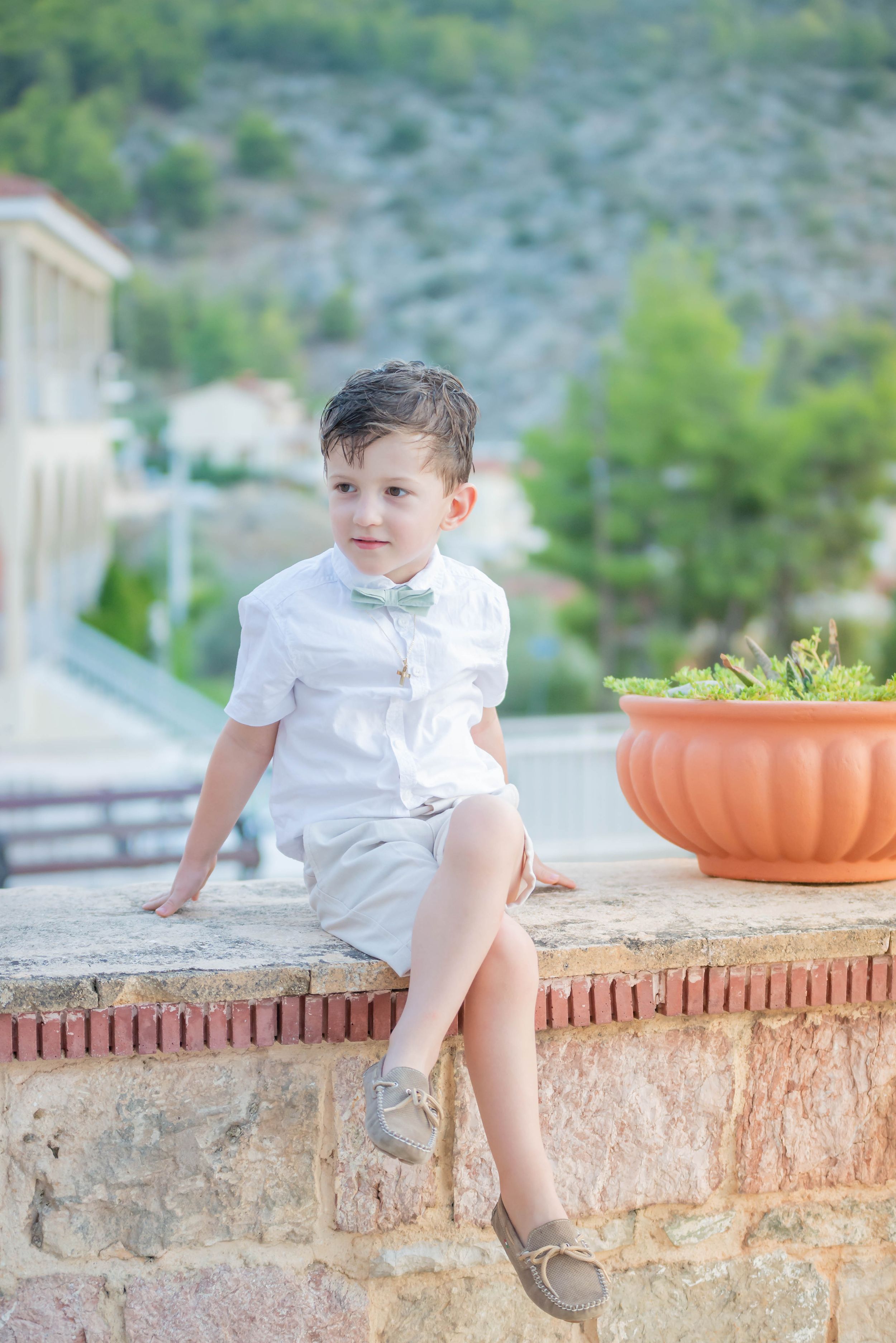 A young boy in a white short-sleeved shirt and light-colored shorts sits on a stone wall outdoors, wearing a light green bow tie.
