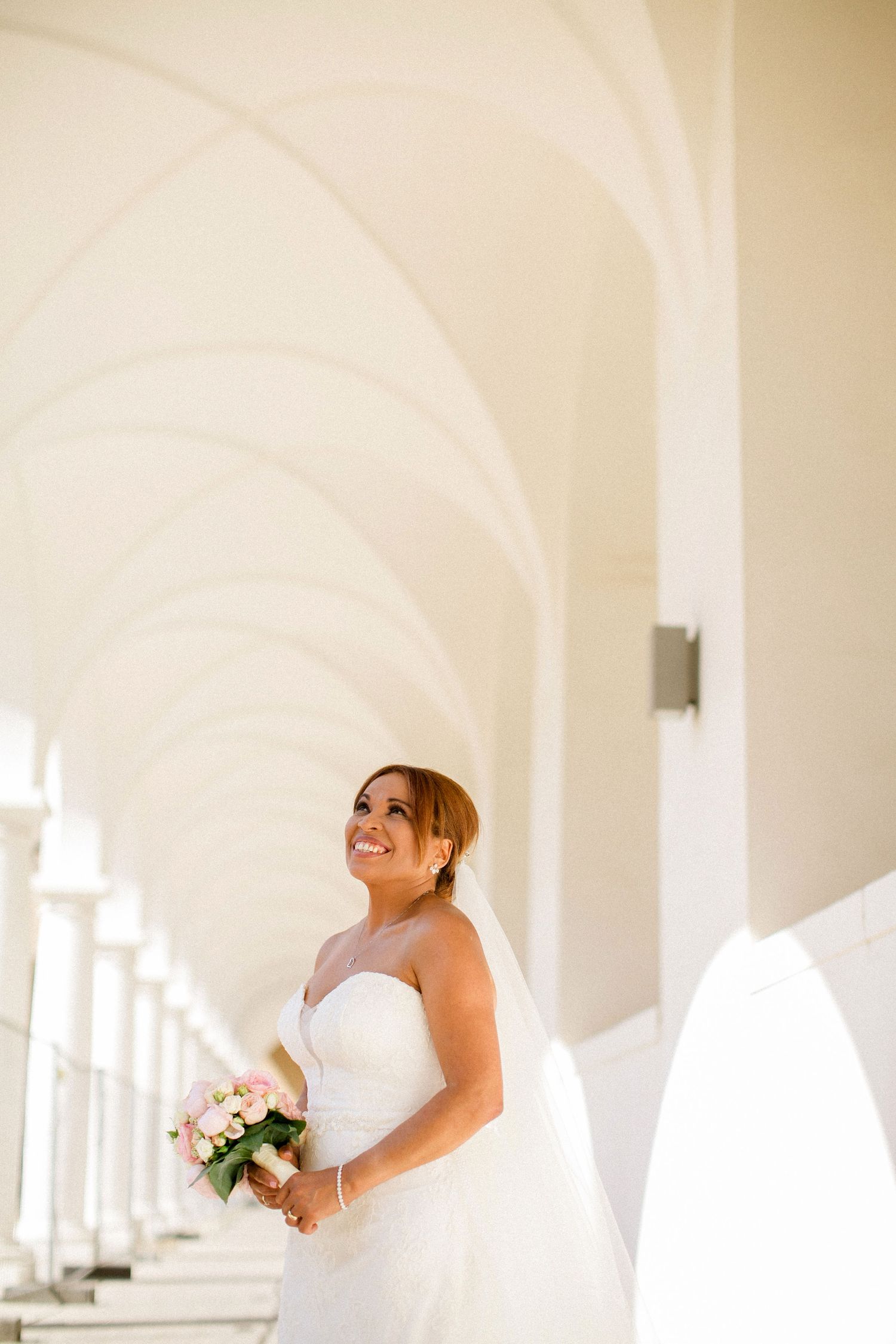 A bride in a white wedding gown holds a bouquet while standing in an elegant white arched corridor.