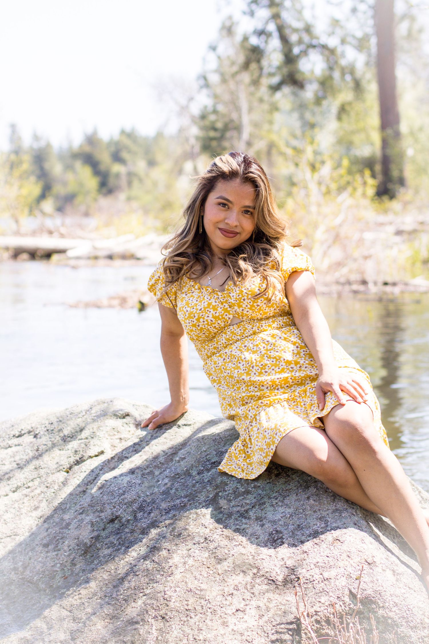 Yellow floral sundress worn on a bright sunny day by a lake with rocks and evergreen trees in the background.