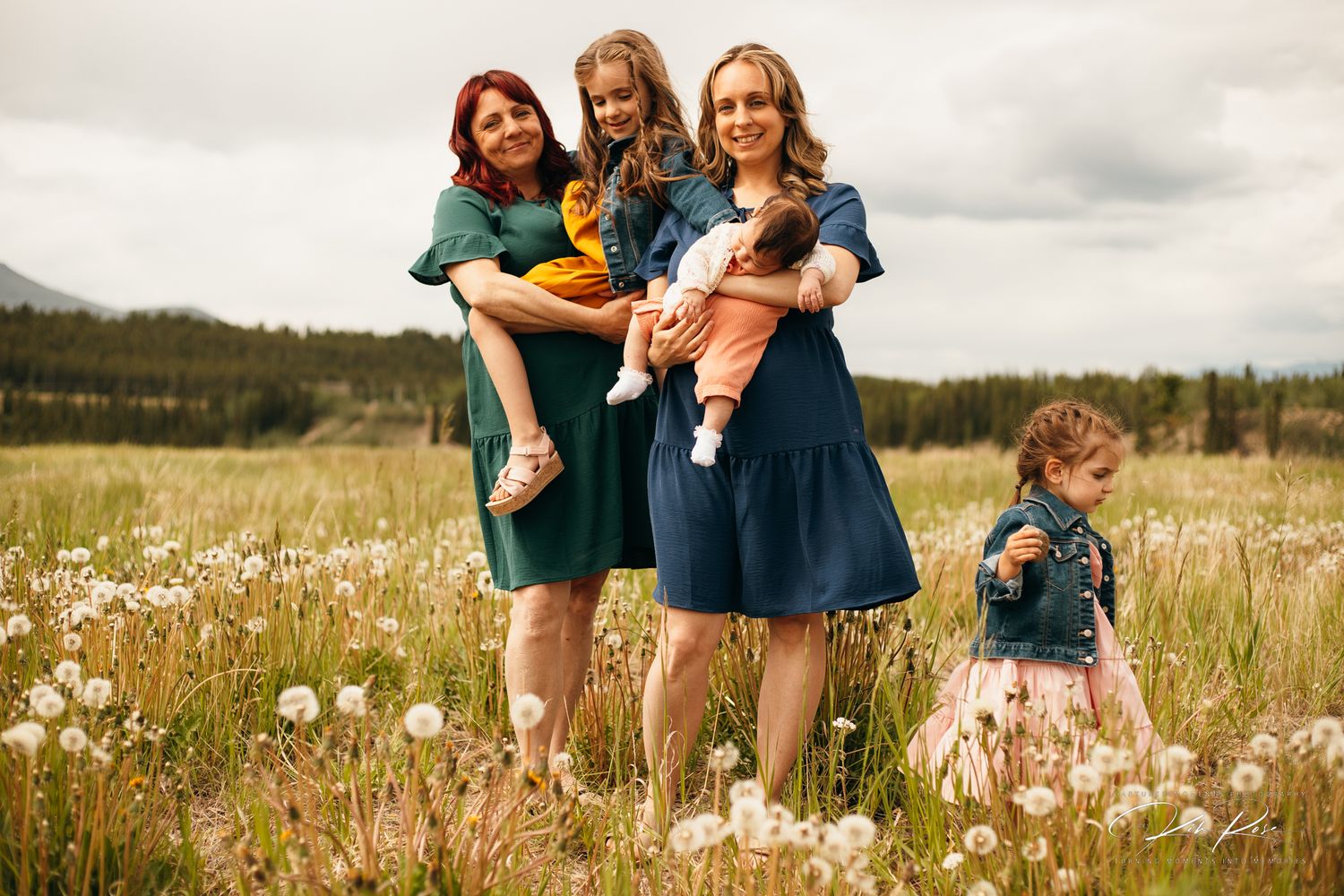 Two women holding children posing in a dandelion field with mountains behind them.