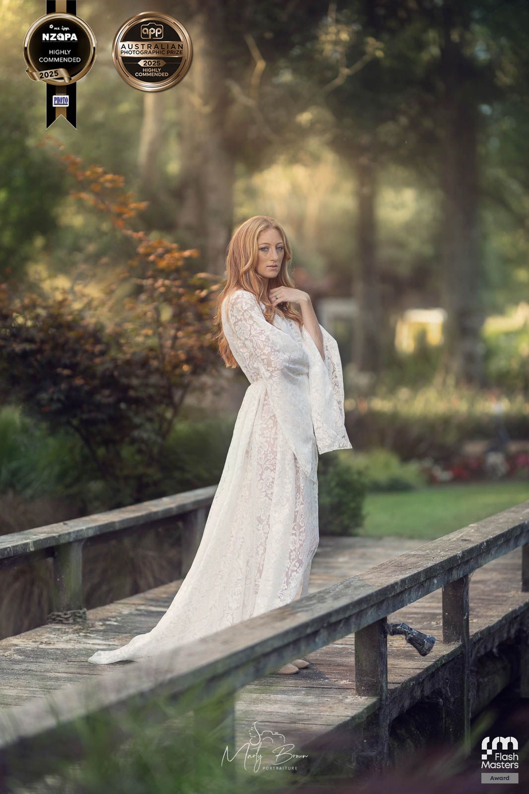 Girl in long white flowy dress standing on wooden bridge with warm sunset light illuminating the trees behind.