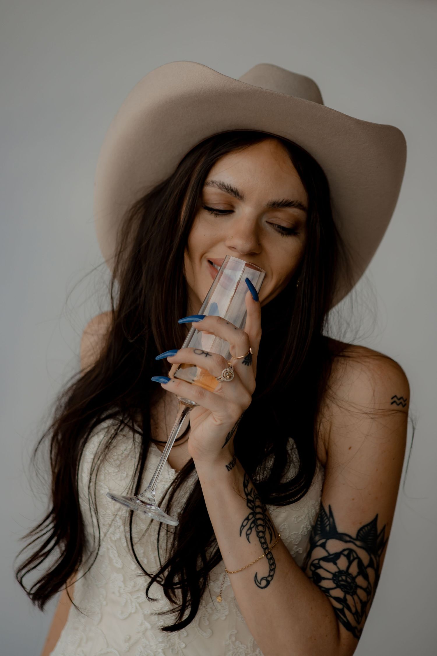 A bohemian bride in a cowboy hat sips champagne while showing off her arm tattoos.