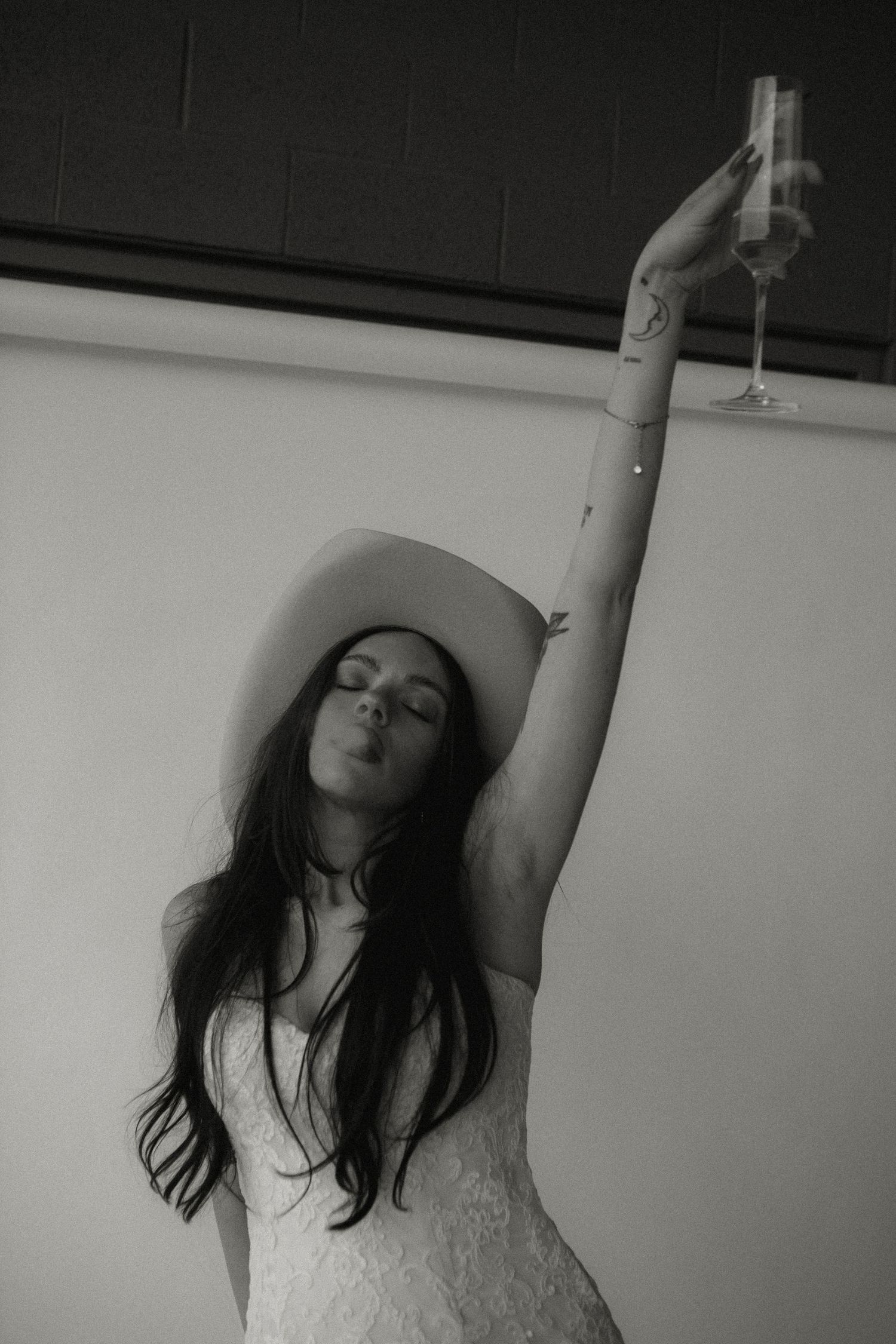 Moody black and white photo of bride in cowboy hat raising champagne glass.