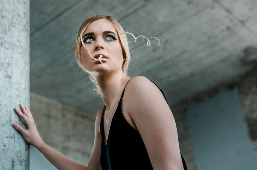 Young blonde woman in black dress leaning against concrete pillar looking upward dramatically.