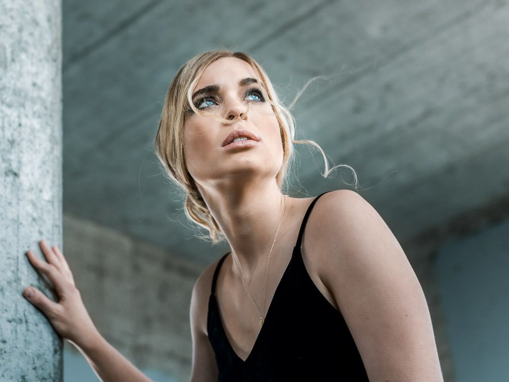 Blonde woman in black dress leaning against concrete wall gazing upward in urban setting.