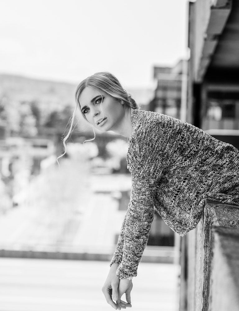 Black and white photo of a blonde woman in knit sweater leaning on a railing outdoors.