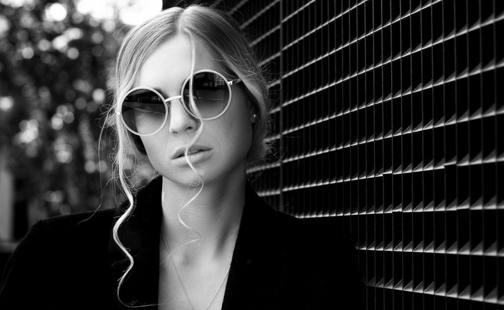 Black and white portrait of blonde woman wearing round sunglasses against slatted wall.