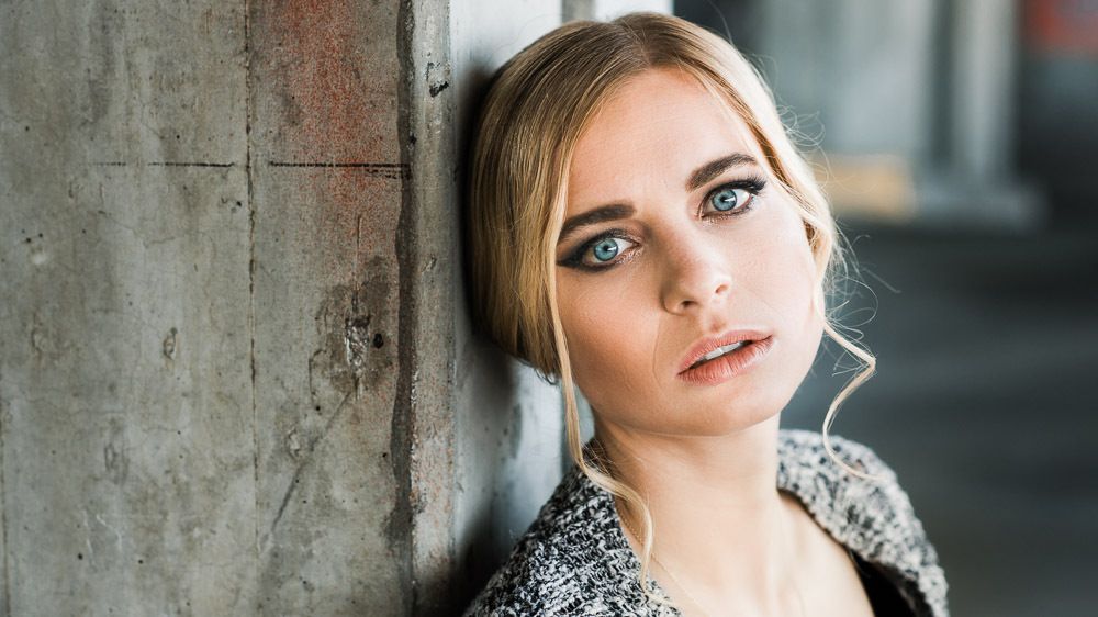 Close-up portrait of a blonde woman with blue eyes and textured knit cardigan against concrete wall.