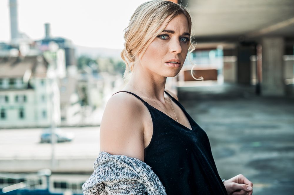 Blonde woman in black dress with off-shoulder knit cardigan posing in an urban outdoor setting.