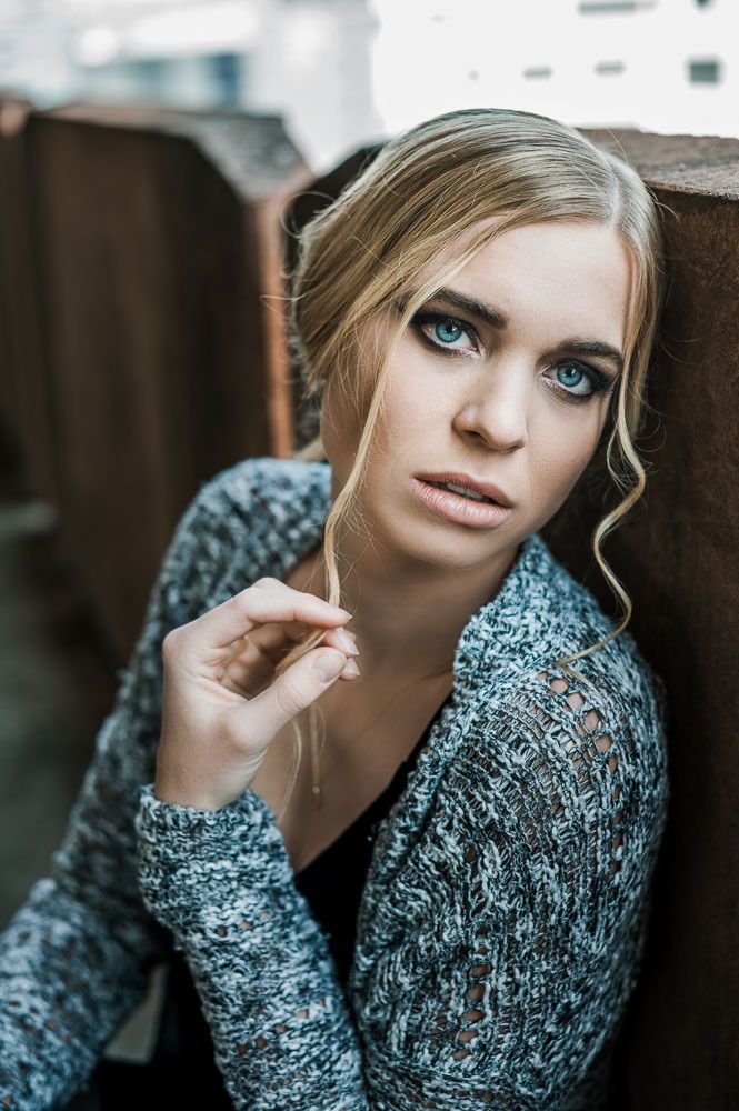 Close-up portrait of a blonde woman with blue eyes in a grey knit cardigan leaning against a wall.