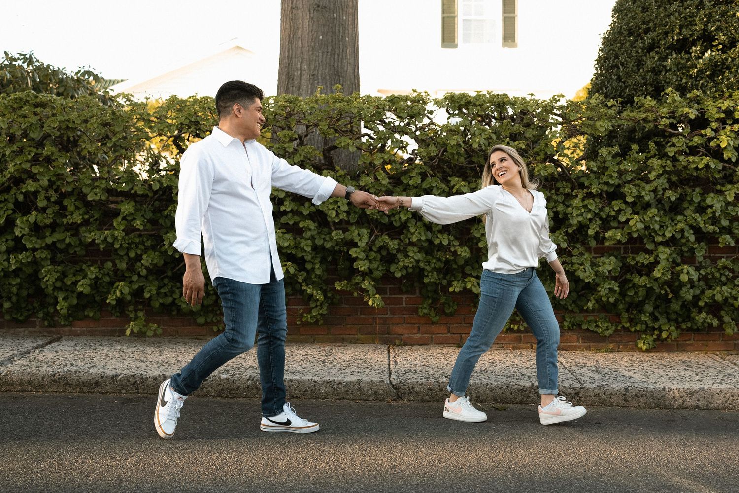 Playful couple holding hands while walking on sidewalk near white picket fence.