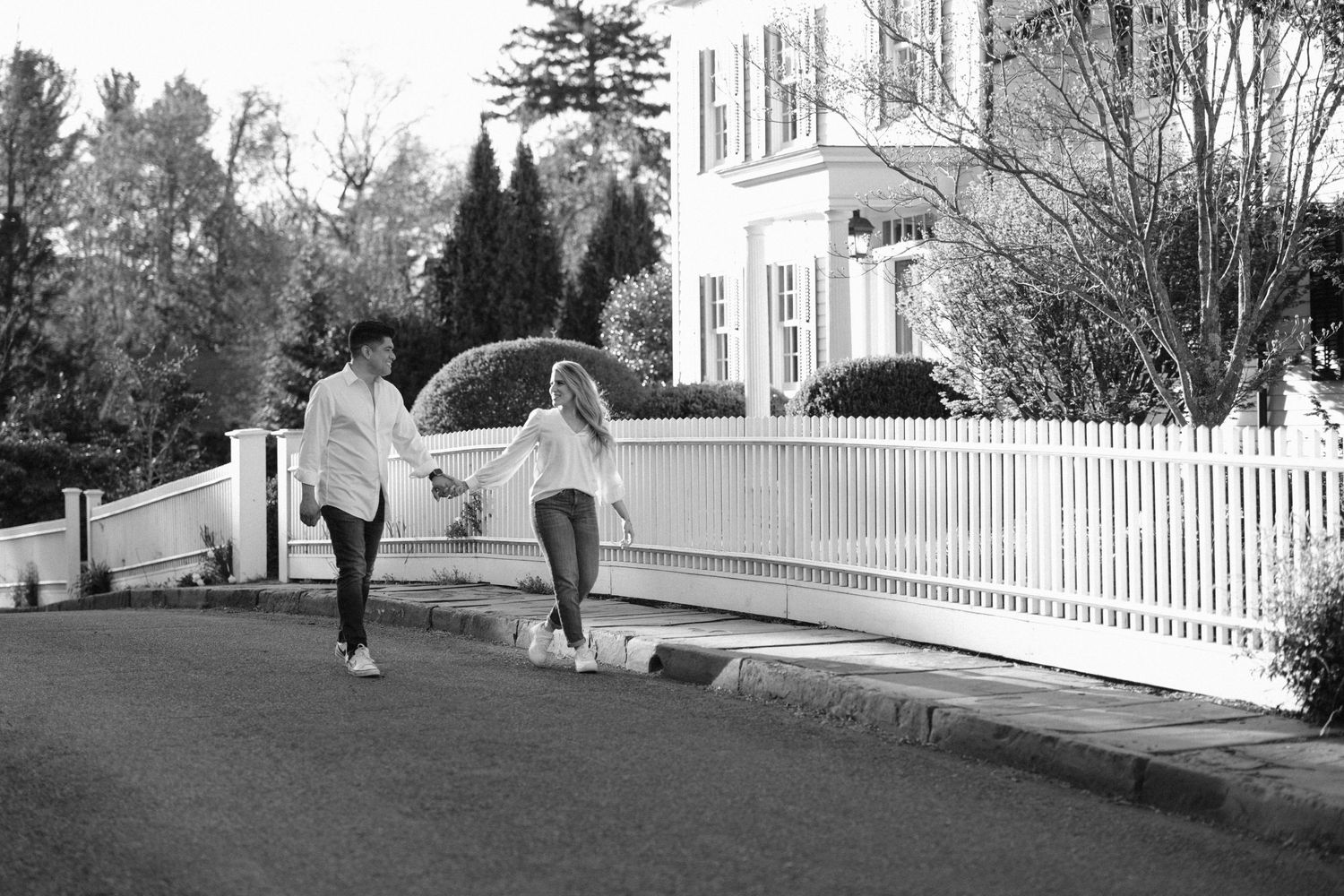 Black and white photo of couple walking together along residential street with white fence.