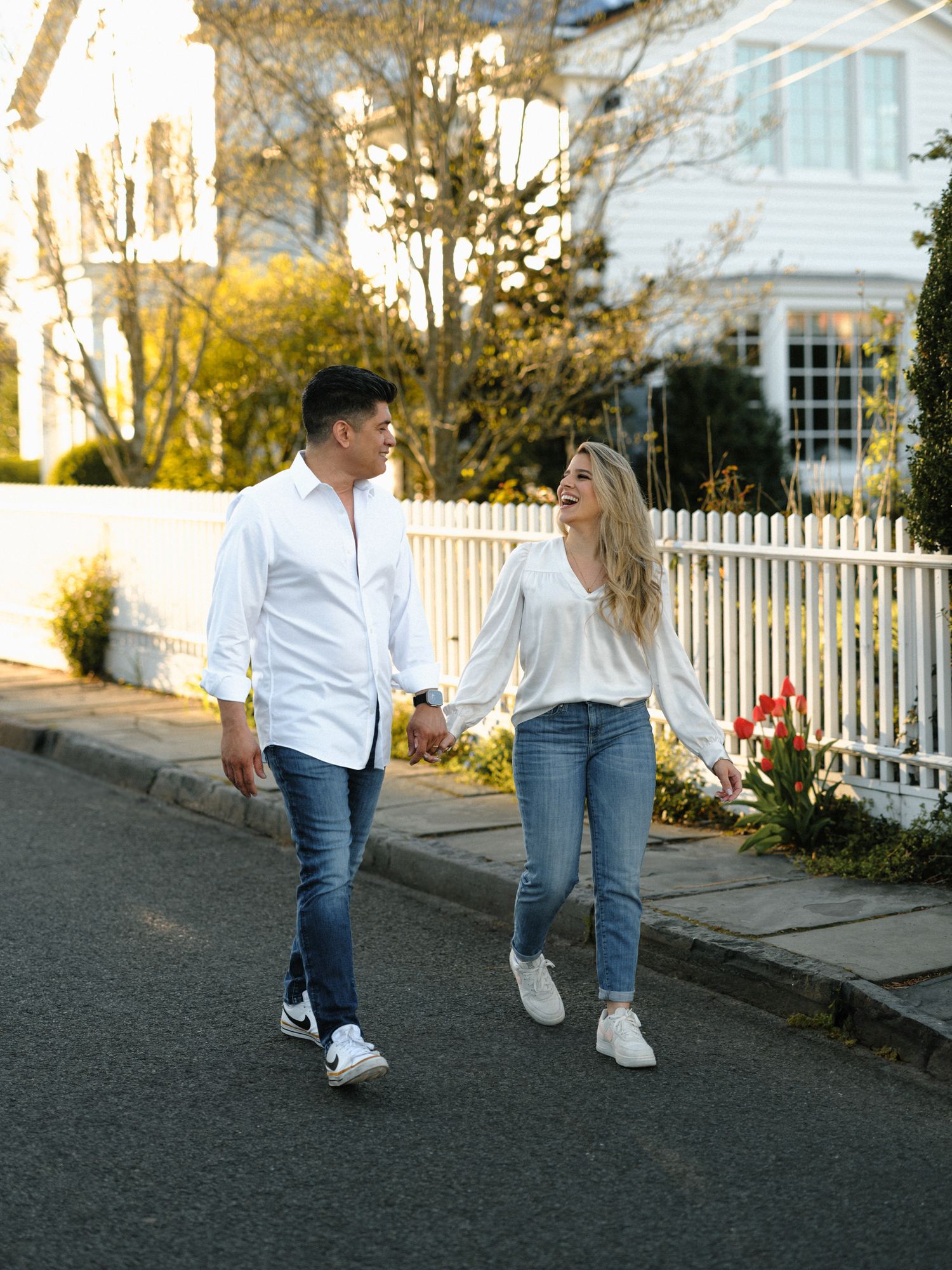 Casual couple walking together on street with white picket fence and classic home in background.