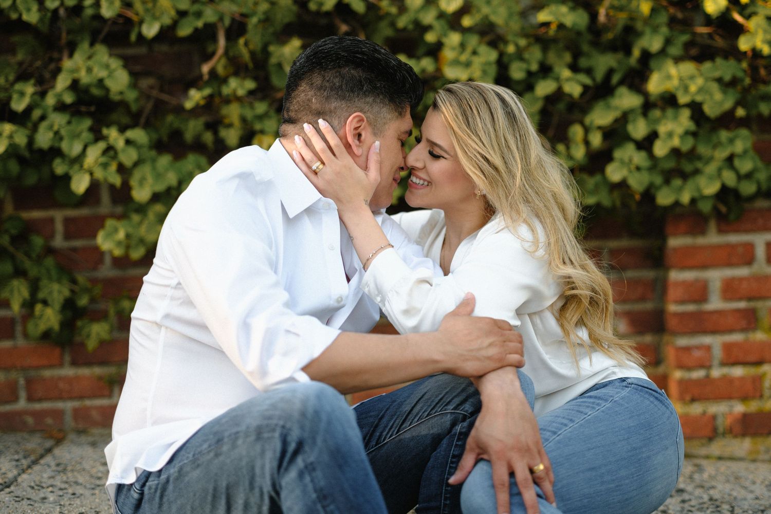 Couple sharing a tender moment while sitting on a curb with greenery in background.
