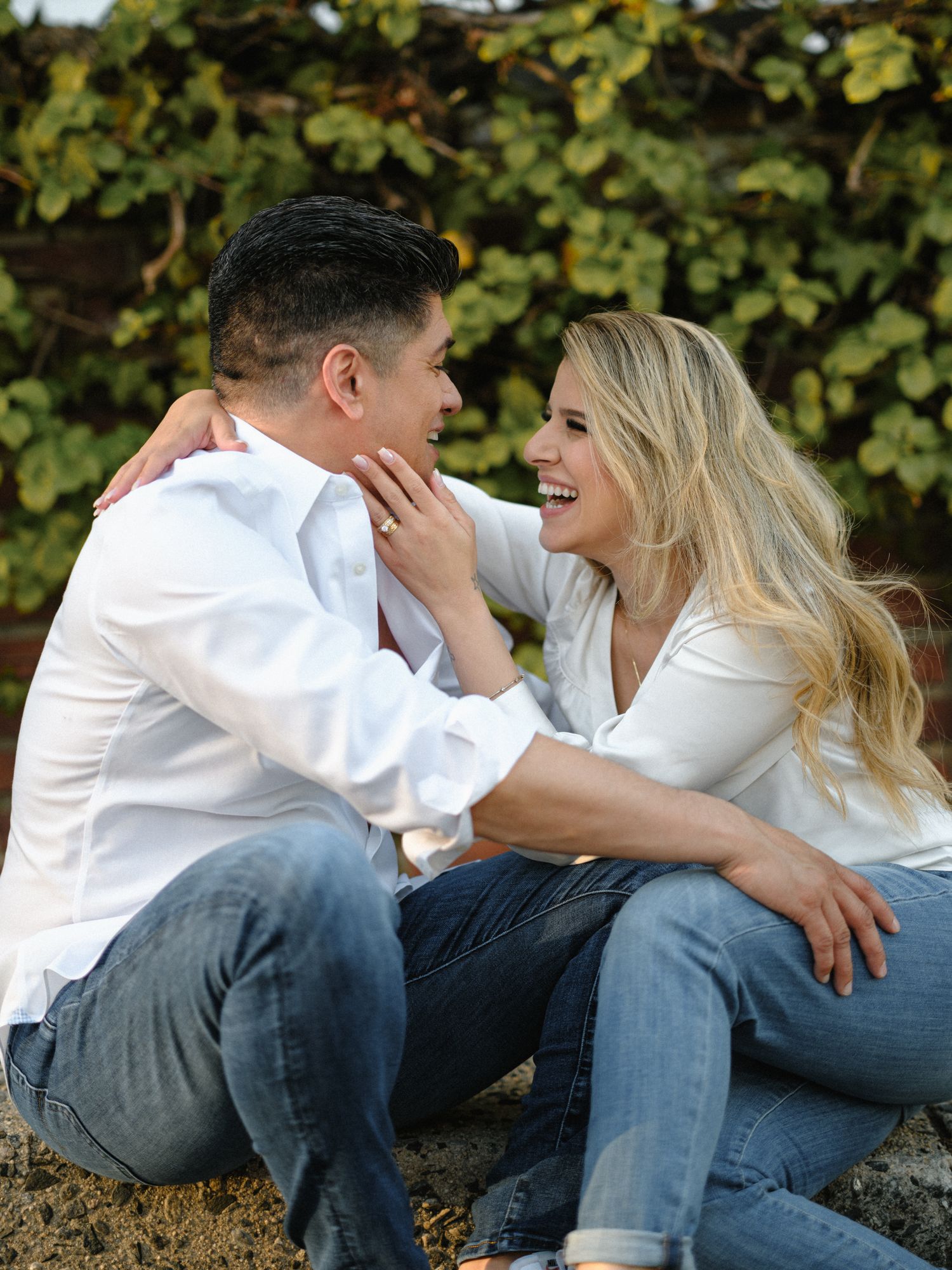 Couple laughing together while sitting on curb against leafy backdrop.
