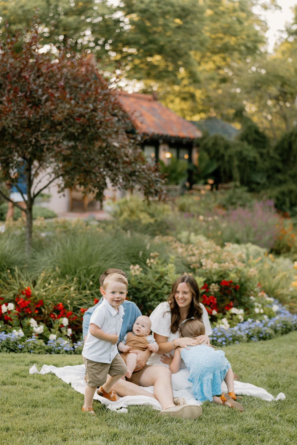 Family enjoying a sunset picnic together on a blanket in a lush garden with red flowers and greenery in the background.