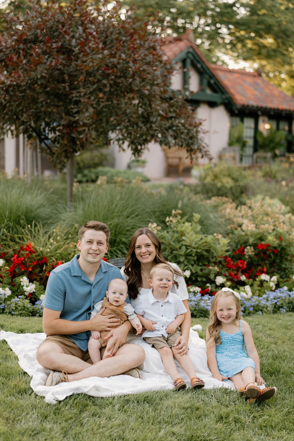 Family relaxing together on white blanket in garden with red flowers and greenery in background.