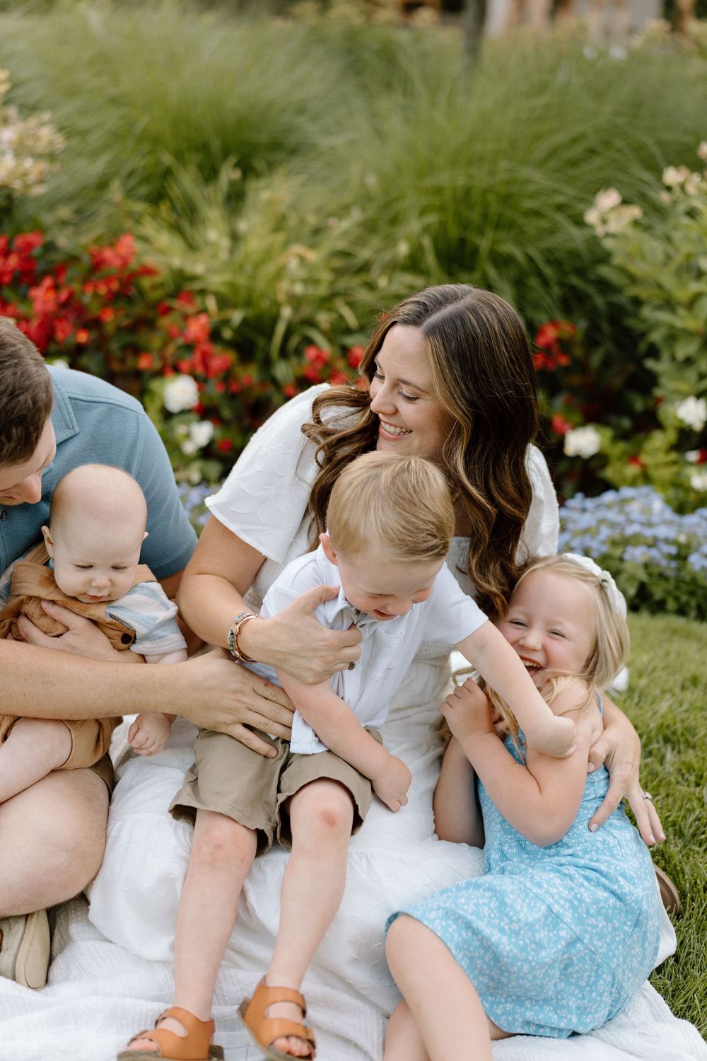 A family in white clothing sits together in a garden with blooming flowers and greenery in the background.